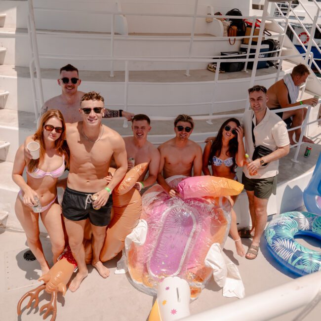 A group of friends stand on a boat deck, smiling and holding inflatable pool toys. They are dressed in swimwear, enjoying the radiant sunshine. In the background, there are more people and part of the boat's structure, creating a vibrant and fun atmosphere perfect for sailing adventures.