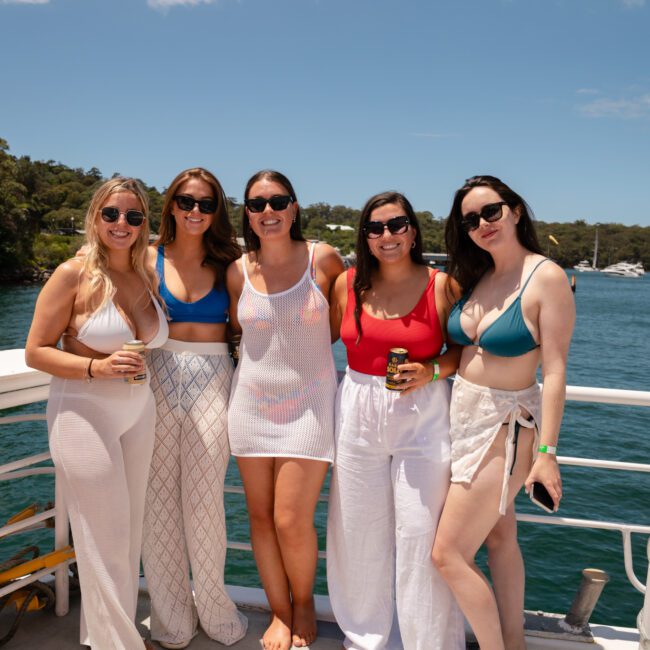 Five women dressed in summer attire stand on the deck of a boat, smiling at the camera. The background features lush green trees and bright blue water under a clear sky, creating a relaxed, vacation-like vibe.
