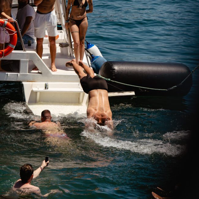 A group of people are enjoying a day on a boat. One person is diving into the water from the back of the boat, while others watch or swim nearby. A large inflatable raft is floating close to the boat with logos in image corners saying "Sail Gustavo" and "The Yacht Social Club." Enjoying carefree moments at sea!