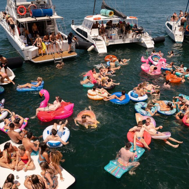 A lively boat party with many people floating on inflatable pool toys in the water, surrounded by anchored yachts. Some inflatables are shaped like swans, unicorns, and flamingos. People are socializing, sunbathing on paddleboards, and enjoying the festive atmosphere.