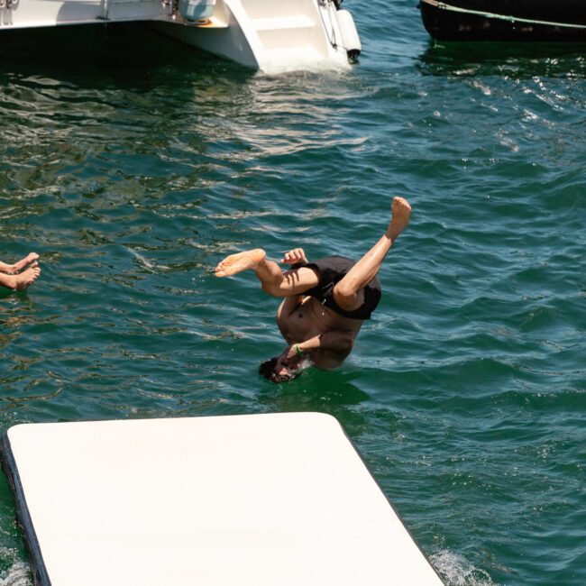A person is mid-flip in the air, about to enter the water near a boat. Shirtless and wearing black swim trunks, they appear to be diving off a floating mat. Other people can be seen on the boat and a nearby float in the vibrant blue-green water.