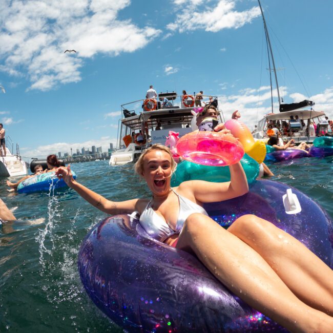 A woman in a white bikini smiles and waves while floating on a purple inflatable ring amidst a group of people on colorful inflatables in the water. Behind her, catamarans with more people sail under a blue sky with scattered clouds, all enjoying the picturesque scene.