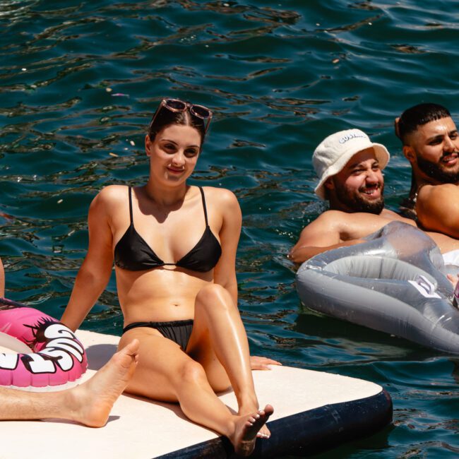 A group of people enjoy a sunny day at the beach. Two individuals relax on a floating platform, while others lounge on inflatable pool toys. All are in swimwear, with clear blue water visible in the background.