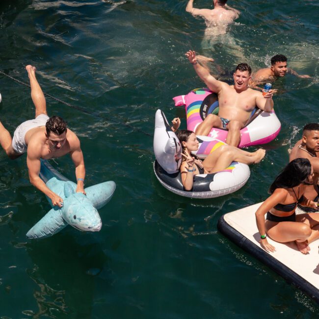 A group of people enjoys a sunny day on the water. Some are floating on inflatables, including a shark and a flamingo, while others are in the water playing with paddleboards. Everyone is smiling, holding drinks, and wearing swimsuits. A boat and a floating deck are also visible.