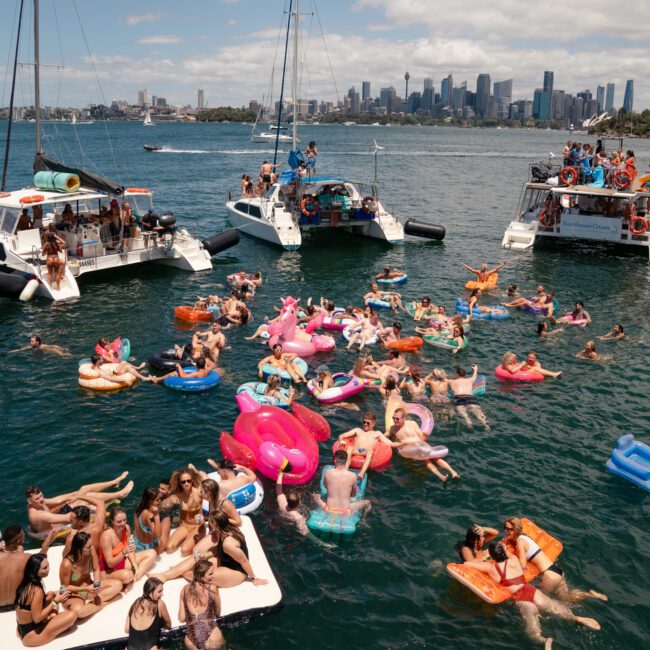 A large group of people enjoy a sunny day on a lake, floating on colorful inflatables. Four boats are moored nearby, and the city's skyline is visible in the background. The mood is lively with many people socializing, swimming, and relaxing to music on the water.