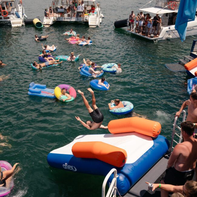 A lively scene on a sunny day features people swimming and floating on inflatables between several docked boats. Some are lounging on the boats, while others jump into the water. A bridge and city skyline enhance the picturesque background.