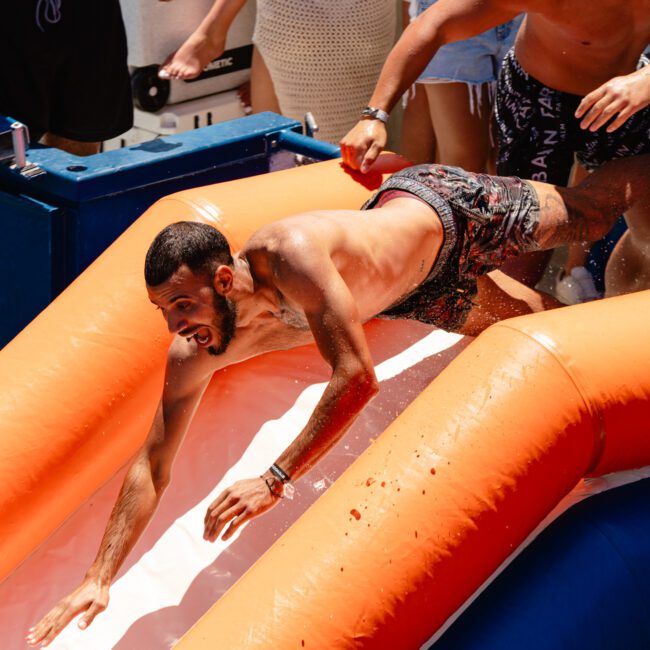 A man wearing swimming trunks slides headfirst down an orange inflatable water slide with an enthusiastic expression, both arms extended forward. The background reveals another person in swimwear and an outdoor setting, suggesting a joyful, sunny environment perfect for some family fun.
