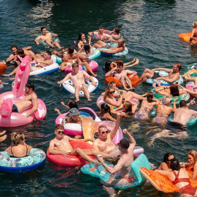 A lively group of people enjoy a sunny day on a body of water, lounging on colorful inflatable floaties shaped like animals, fruits, and other fun designs. The scene is festive and vibrant, with many people holding drinks and chatting under the bright sun.