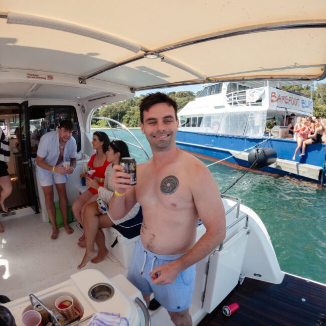 A group of friends on a boat enjoying a sunny day. The man in the foreground, wearing swim trunks, is smiling and holding a beverage can. Several others are seated or standing behind him, also with drinks in their hands. Another boat filled with happy passengers is visible in the background.