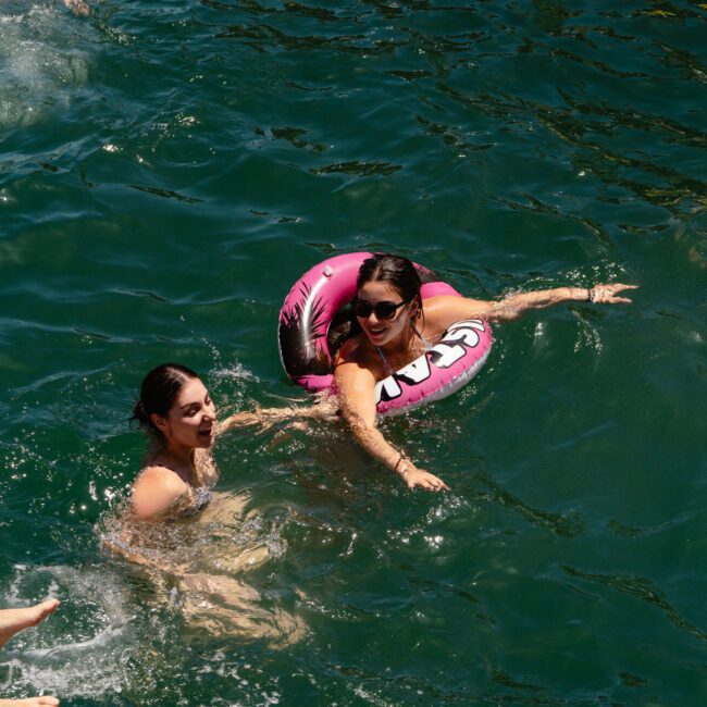 Two women are swimming in a body of water. One woman is wearing sunglasses and relaxing with a pink donut-shaped float. They appear to be enjoying a bright, sunny day. The image features logos in the bottom corners: "SAL GUSTAVO" on the left and "Light Social" on the right, giving it a vibrant feel.