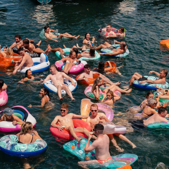 A large group of people enjoys a sunny day in a lake, floating on various colorful inflatables. Some are on flamingo and pizza-shaped floaties, among others. Everyone appears to be relaxed and having fun, with boats cruising in the background.
