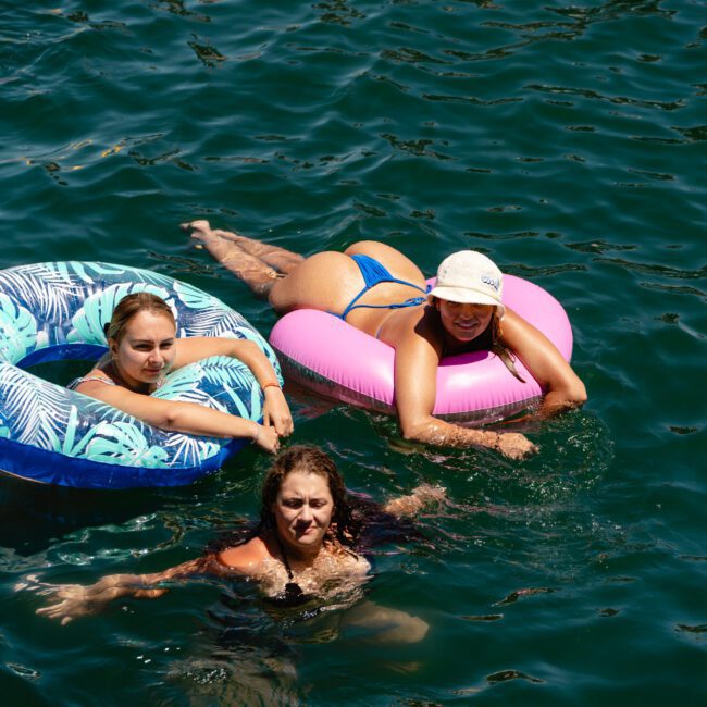 Three individuals enjoying the water: two in inflatable rings, one blue and the other pink, while the third is swimming. They appear relaxed and smiling under a bright, sunny sky. Logos in the bottom corners read "Sal Gustavo" and "Yacht Social Club," highlighting a perfect day on the water.