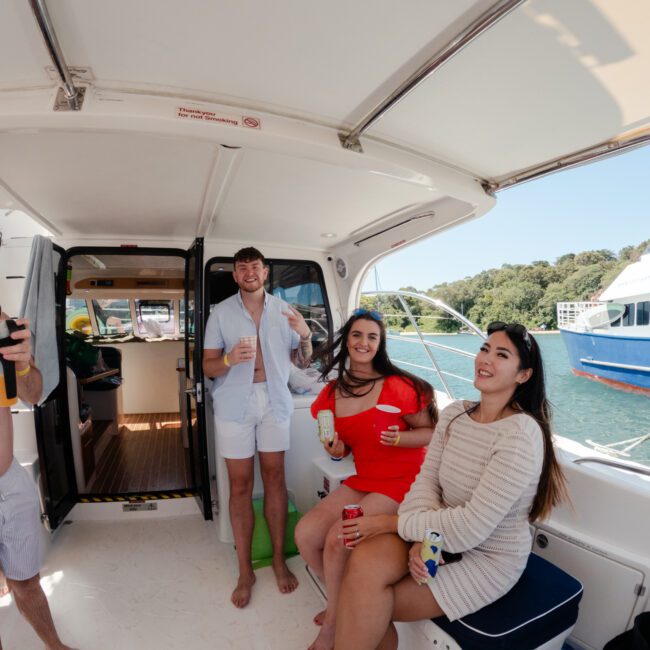 A group of people enjoying a sunny day on a boat. A man on the left is opening a bottle, while others are sitting and standing around, holding drinks and smiling. The boat's cabin is visible in the background, with another sailboat seen on the water behind them.
