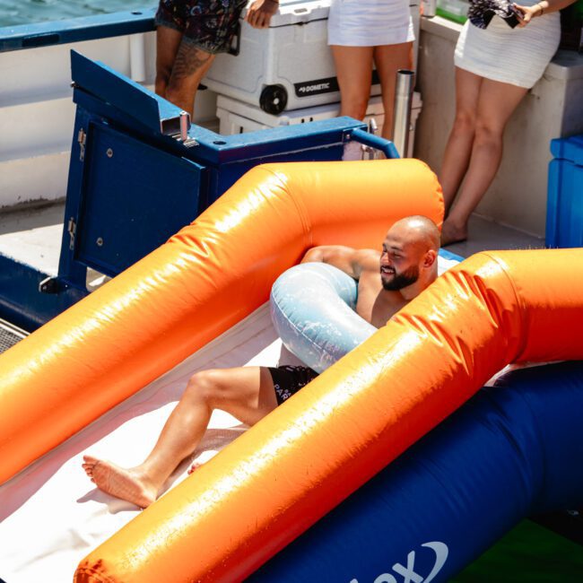 A person slides down an orange inflatable slide into the water, holding a light blue pool float. Others on the boat deck watch, some holding drinks. The scene is sunny and cheerful, with people dressed in swimwear. Logos of "Sal Gustavo" and "Yacht Social Club" add to the festive atmosphere.