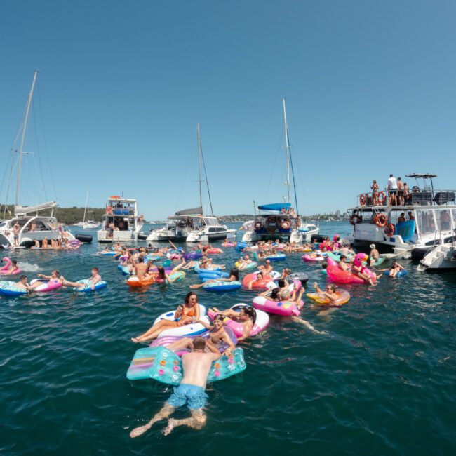 A lively scene of people enjoying a sunny day on the water, floating on colorful inflatable rafts near a cluster of boats and yachts. The sky is clear and the atmosphere is joyful, with many participants reveling in the festivities and basking in the serene weather.
