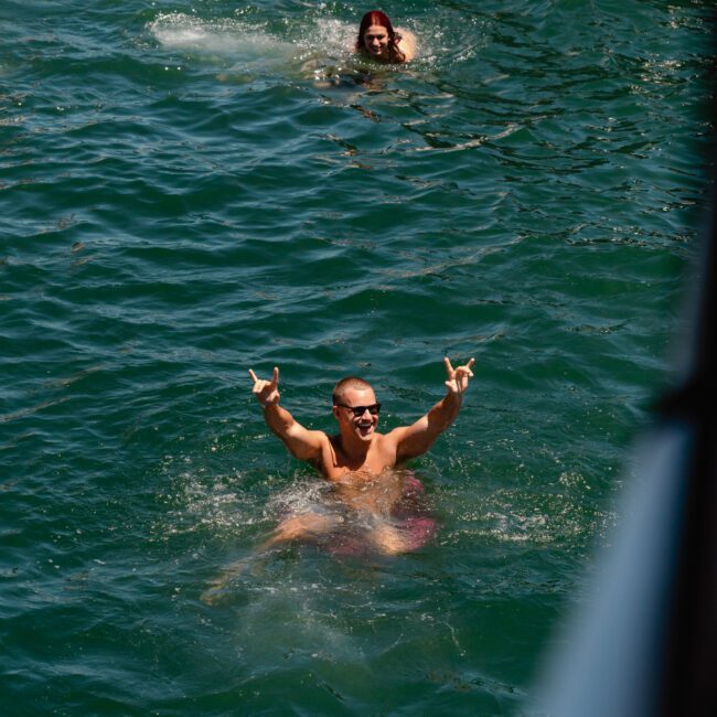 Two people enjoying a refreshing swim in the ocean near the side of a boat. The closer individual, shirtless and wearing sunglasses, makes a celebratory hand gesture with both hands raised high. The clear blue water surrounds them as logos for "Sail Gustavo" and "Yacht Social Club" are visible in the background.