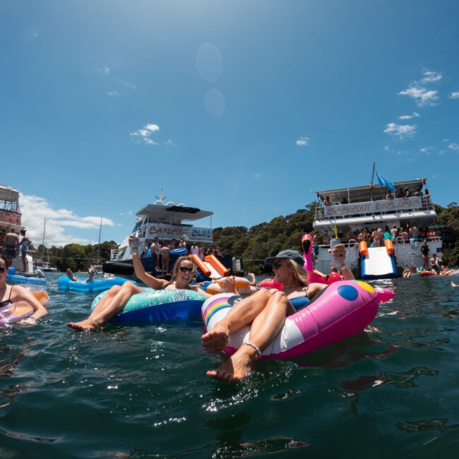 People are floating on inflatable rafts in the water near large boats under a sunny sky. Some are sunbathing while others are swimming or standing on the boats, enjoying a lively atmosphere. The backdrop includes lush green hills and a clear blue sky, creating a picturesque scene.