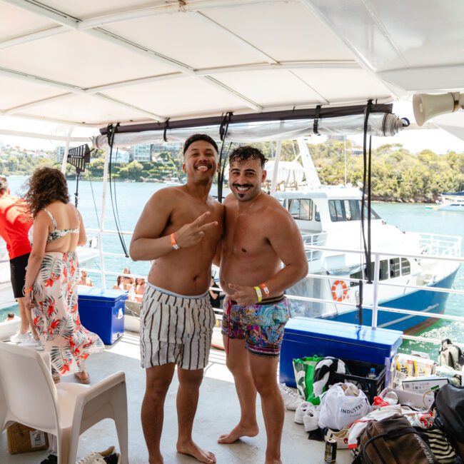 Two smiling men stand on a boat deck, one shirtless and the other in a colorful swimsuit. They're surrounded by scattered bags and coolers. The boat is anchored near a scenic shoreline with trees and water visible in the background, while others enjoy the tranquil setting nearby.