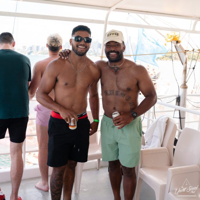 Two shirtless men, smiling on a boat and both holding drinks. One wears sunglasses, a watch, red swim shorts, and a green wristband. The other has on a cap and light green swim shorts, showcasing tattoos on his chest and stomach. Other people are visible in the background enjoying the sunny day.