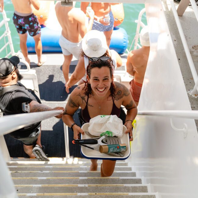 A smiling person with wet hair in a swimsuit carries a tray with food and drinks while climbing the stairs of a boat. Other people in swimwear enjoy the sunny day, some on the lower deck near an inflatable water mat, creating a festive atmosphere on the water.