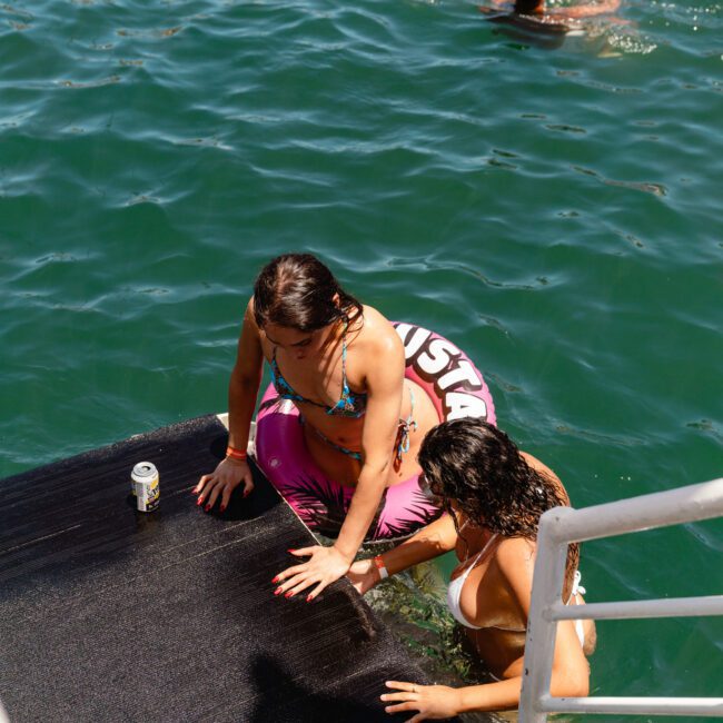 Two women in swimsuits climb onto a black platform from the water. One woman holds a pool float with the word "Gustavo" visible. A drink can rests on the platform beside them. Other swimmers enjoy the surrounding water. Text on the image reads "SAIL GUSTAVO," capturing a lively scene of summer fun and adventure.