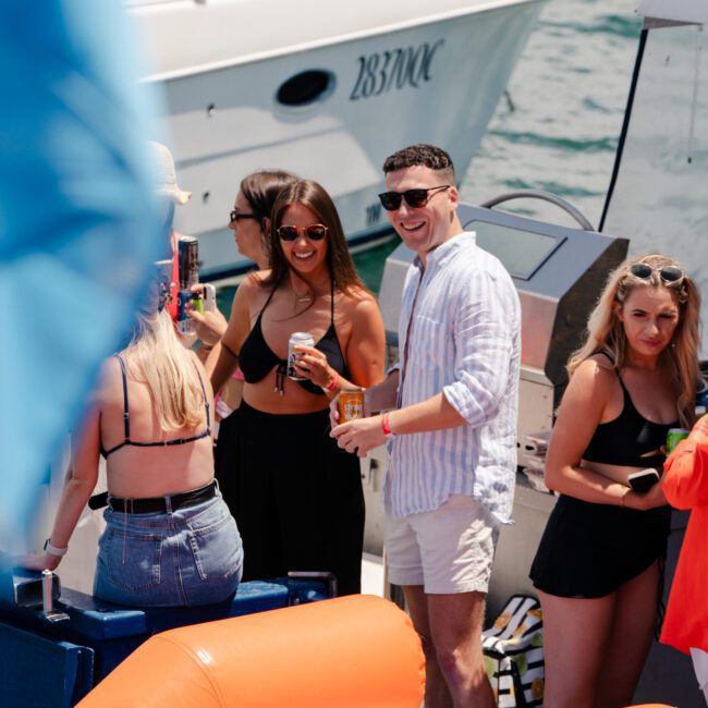 A group of people enjoys a sunny day on a boat. They are casually dressed, with some wearing swimsuits. Two individuals, a woman in a black bikini top and a man in a striped shirt, laugh and hold drinks. An orange inflatable ring is in the foreground, adding to the cheerful atmosphere.