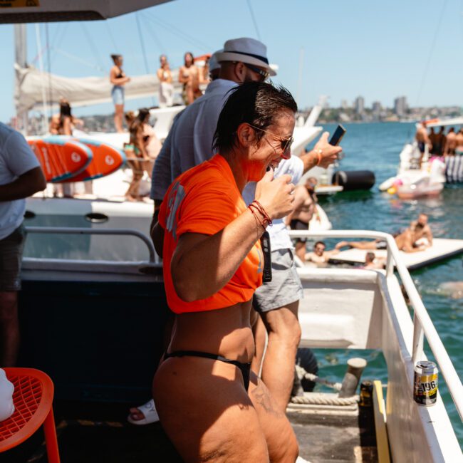 A woman in a vibrant orange shirt and dark shorts is joyfully dancing on a boat deck, surrounded by people. The boat sails on a sunny day out at sea, with other boats and cheerful folks in the background. A can of drink rests on a nearby ledge. The festive event is lively and spirited.