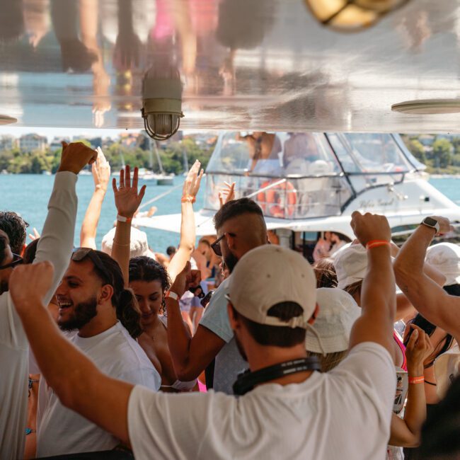 A lively group of people aboard a boat are dancing and raising their hands in celebration. The boat is docked near the water with other yachts visible in the background. The sky is clear and sunny, adding to the festive atmosphere. Logos reading "Sail Gustavo" and "Lose Control Yacht" are present.