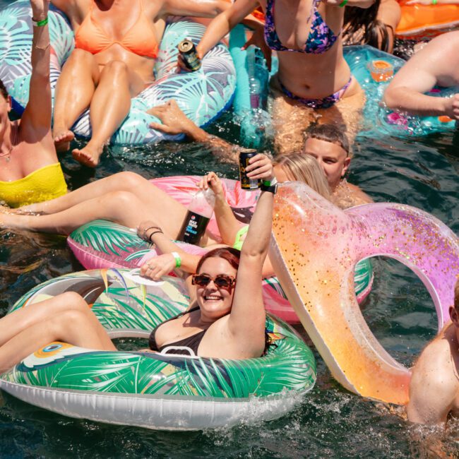 A group of people enjoying themselves on colorful inflatable unicorn pool floats in a body of water. They are smiling, laughing, and holding drinks, creating a festive and lively atmosphere under the sun.