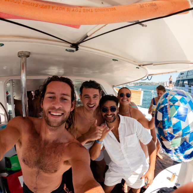 A group of four friends takes a selfie on a boat under a shaded canopy. One person, holding the camera, has long hair and is smiling broadly. Two men, one with a thumbs-up, and a woman wearing sunglasses stand alongside. A vibrant birthday balloon floats beside them, adding to the celebration atmosphere.