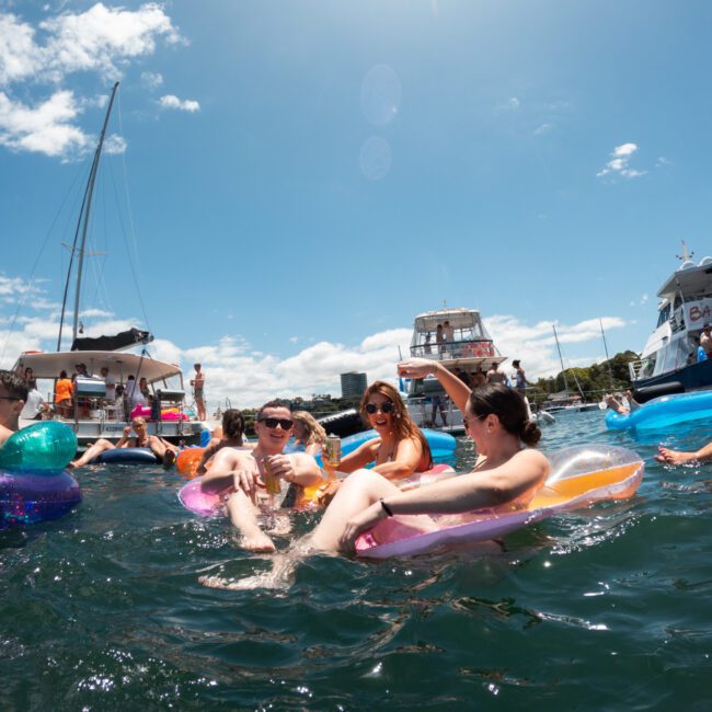 People relaxing and socializing on colorful floaties in the water with sailboats and yachts in the background on a sunny day. The scene is lively with clear blue skies and vibrant water, featuring individuals enjoying drinks and floating near an anchored catamaran.