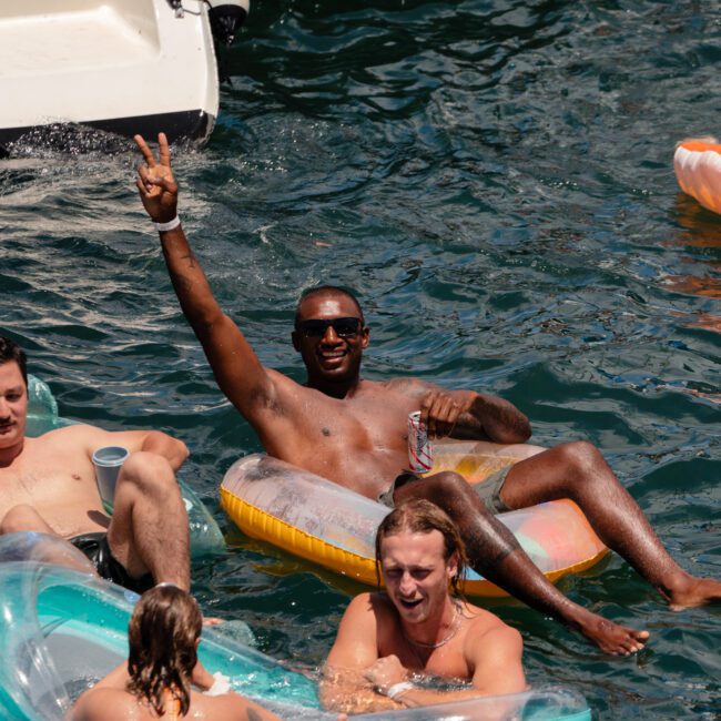 A group of people are enjoying a sunny day in the water next to a boat. One man in an inflatable float holds a can and raises his hand giving a peace sign. Others are also floating and socializing, creating an inviting atmosphere on the water.
