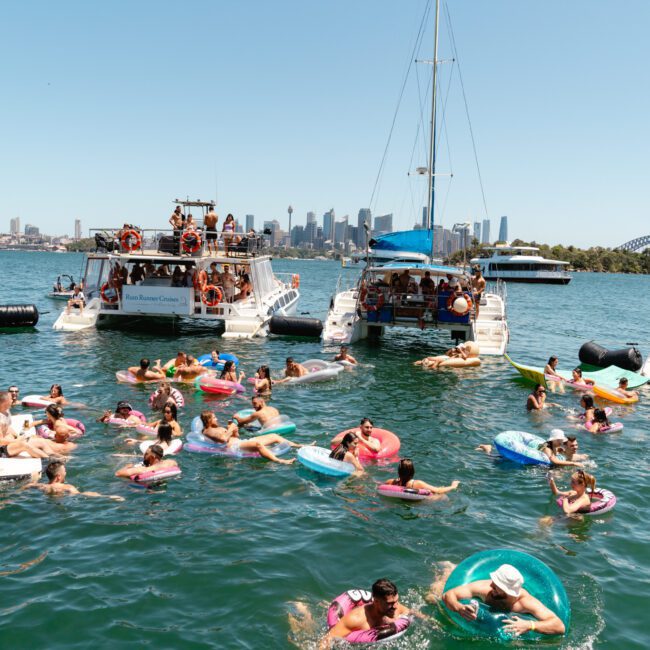 People enjoy a sunny day on the water as they float on colorful inflatables and swim near anchored boats. The boats have passengers on board watching the scene, with a city skyline and iconic bridge visible in the background, adding to the idyllic atmosphere.