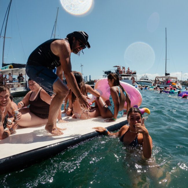 A group of people in swimwear enjoying a sunny day on the water. They are gathered on a floating dock, laughing and smiling, with boats and other watercraft in the background. A person is in the water holding onto the dock, while others sit or stand on it.