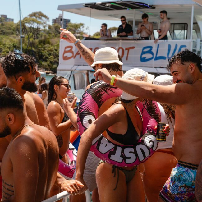 A lively group in swimwear is enjoying a boat party. Some are dancing and holding drinks, while others socialize under clear skies and lush greenery. The boat, named "BLUORT BLUE," prominently features the "Sail Gustavo" and "White Society" logos.