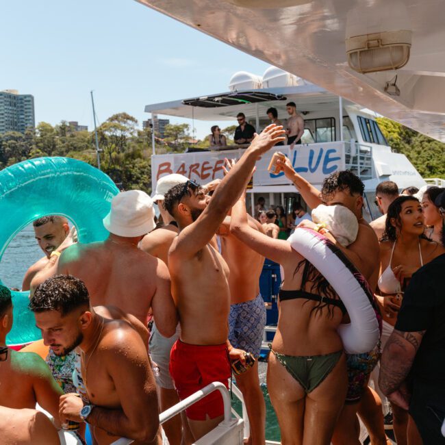 A lively crowd of people enjoy a party on a boat. Some are gathered on a second boat in the background with a "Barely Blue" sign. Attendees wear swimwear, holding inflatables and drinks. Trees and city buildings can be seen in the distance, with the "Sail Gustavo" logo prominently displayed.
