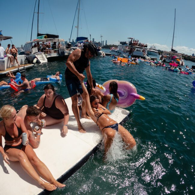 A group of people enjoys a sunny day on the water. Some sit on a floating platform, chatting and relaxing with drinks, while a man helps a woman climb aboard. Several boats and people on inflatables create a lively and festive atmosphere in the background.