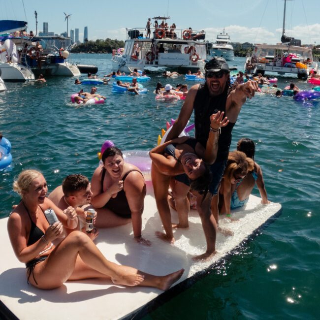 A lively scene of people enjoying a sunny day in the water. Several individuals are on a floating platform, striking playful poses and smiling amidst bustling activity. Others are seen swimming and floating nearby, with boats and stunning city skyscrapers visible in the background.