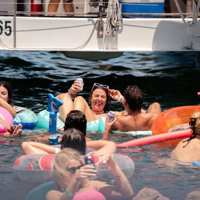 A group of friends are enjoying a sunny day in the water, lounging on various colorful inflatables near a boat. They are smiling, holding drinks, and appear to be having a fun and relaxing time together amidst their laughter and joyful splashes.