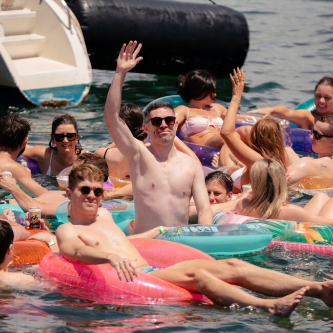 A group of people are floating on inflatable rafts in a body of water, socializing and enjoying the sun. One man in the center stands out, raising his hand triumphantly. Various inflatables in vibrant colors surround them, and a boat is visible in the background.