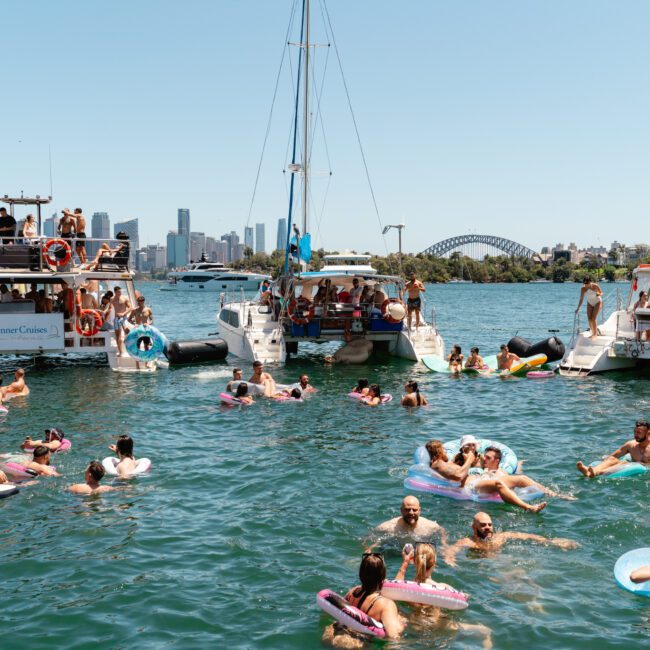Boats are anchored in a body of water dotted with people swimming and relaxing on various inflatables, vibrant with color. A city skyline and a bridge loom in the background. The scene is lively with individuals enjoying a sunny day amidst nature's beauty.