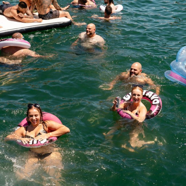 A group of people enjoys a sunny day swimming in a lake beneath the Sail Gustavo and Cath Santos branding. Some float on inflatable tubes, while others swim freely. Two women smile for the camera, holding onto pink inflatables. The atmosphere is lively and relaxed.