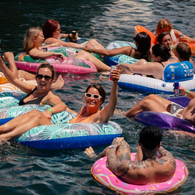 A lively group of people float on inflatable rafts and pool tubes in a body of water, enjoying drinks and the sunny weather. They appear to be having fun, laughing, and relaxing together. The water is calm and everyone is close to each other.