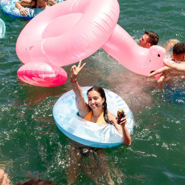 A woman in a blue inflatable ring smiles and gestures a peace sign while holding a can in the water. Around her, other people are also enjoying the water with various inflatables, including a large pink flamingo from "Sail Gustavo" visible in the bottom left.
