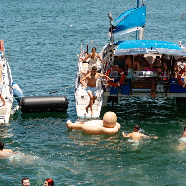People enjoying a sunny day on the water, swimming and relaxing on inflatable rafts near sailboats. The background features a city skyline with tall buildings and lush greenery. Some individuals are sailing, while others are swimming or floating in the refreshing water.