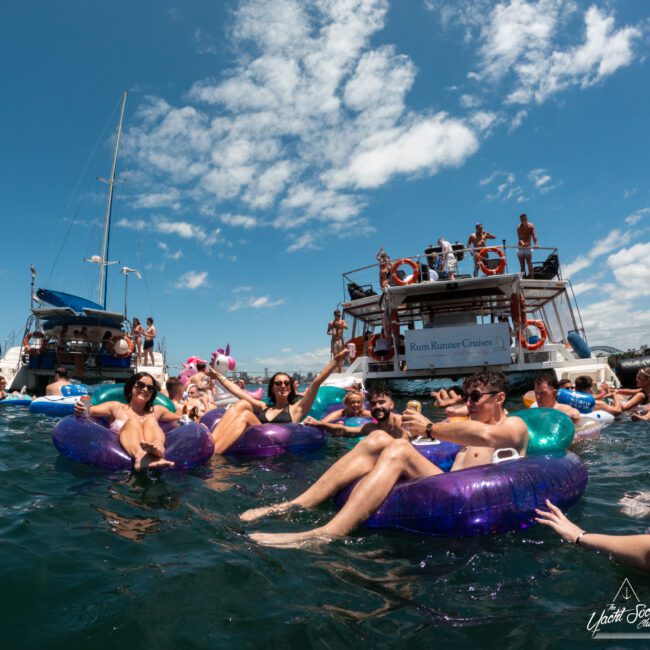 A group of people relax on colorful inflatable pool floats in the ocean, holding drinks and enjoying the sunny weather. In the background, there are two boats with people aboard, and the sky is mostly clear with a few scattered clouds.