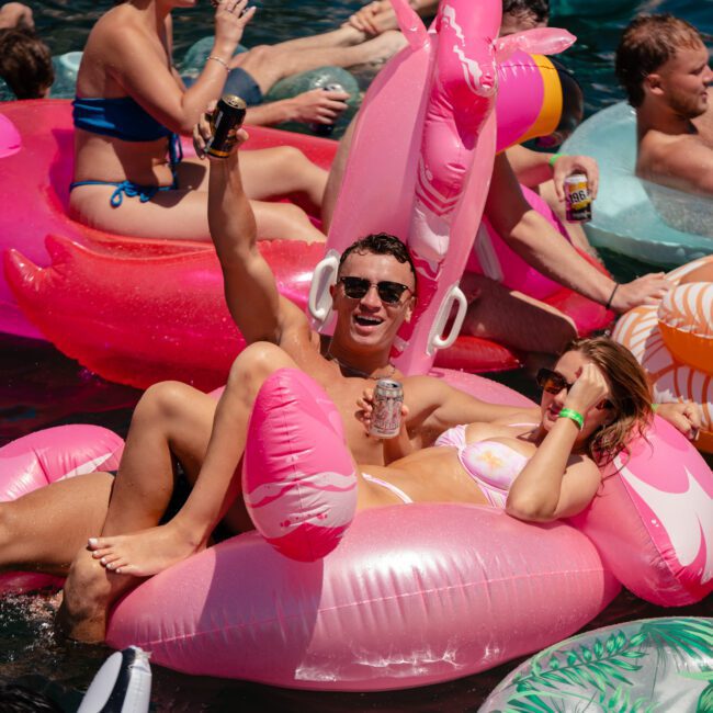 A group of people enjoy a sunny day floating on luxurious inflatable pools in the water. In the foreground, a man raises a can while lying on a large pink flamingo float with a woman beside him. Other revelers are seen on various colorful inflatables in the background, creating a lively atmosphere.