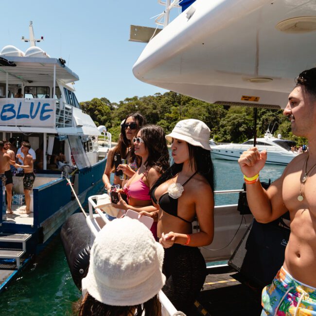 A lively group of people in swimwear enjoy a boat party under sunny skies. Some are on a white yacht, while others are seen on a blue-bordered boat named "Barefoot Blue." Lush greenery is visible in the background, with several kayaks and other boats anchored nearby.