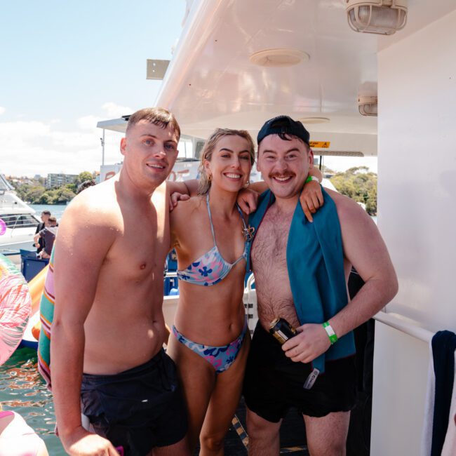 Three people are smiling and posing on a boat under clear skies. A man in swim trunks stands on the left, a woman in a bikini is in the middle, and another man with a drink in hand wearing swim trunks and a life vest stands on the right. In the background, other people and floats dot the water.