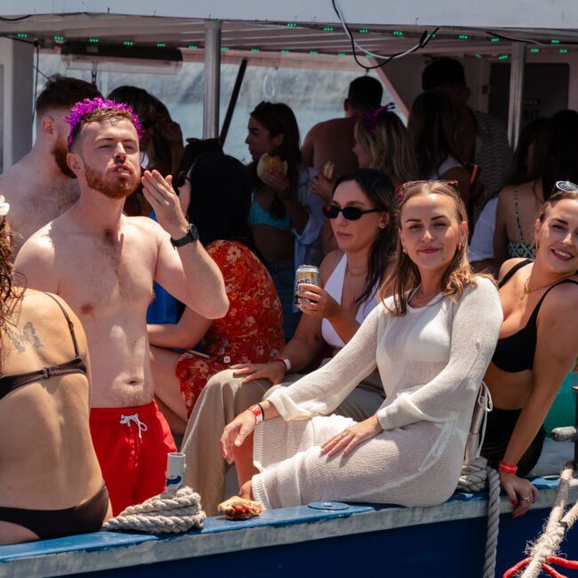 A group of people enjoy a boat party under the sun. Several individuals wear swimwear, while others opt for casual summer clothing. One shirtless man blows a kiss, two women in front pose for the camera, and laughter fills the air as everyone embraces the festive spirit.